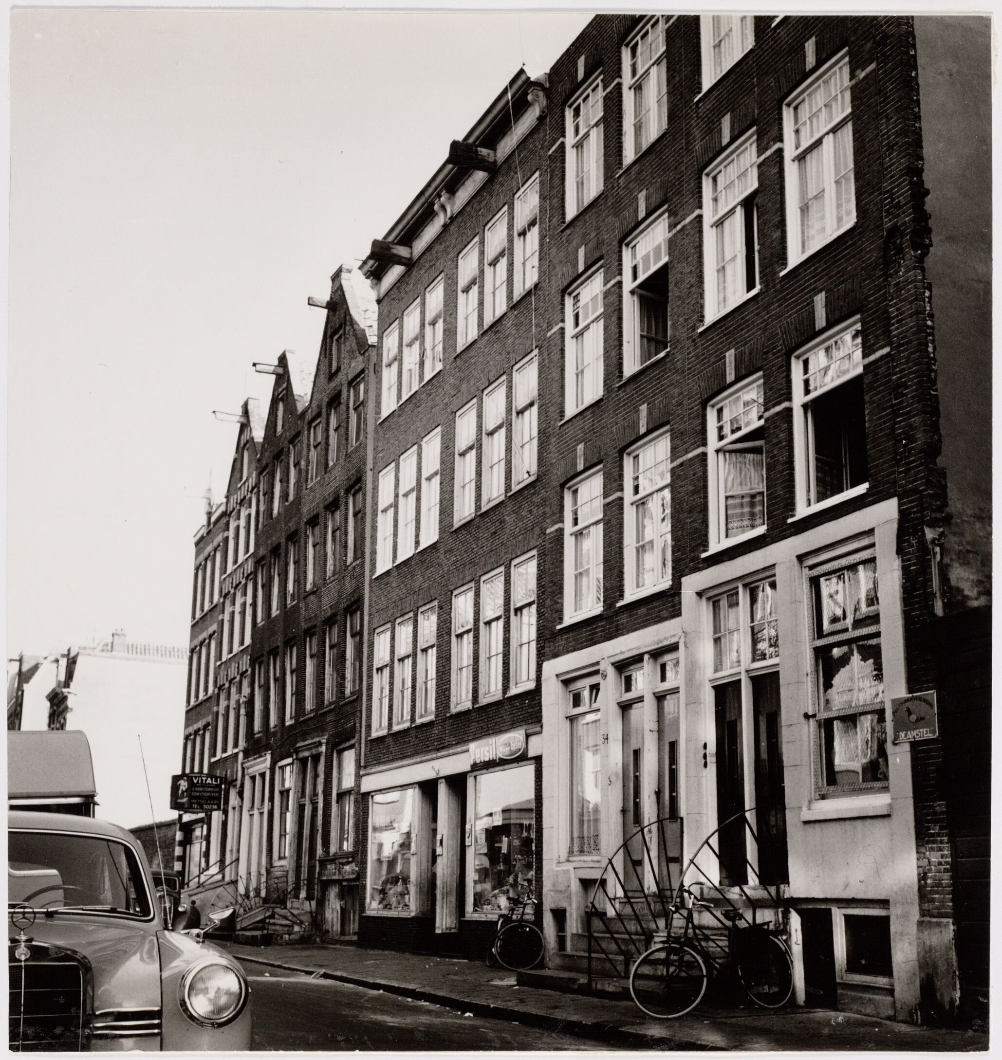 Postwar street photograph of Nieuwe Kerkstraat 32–34 and neighboring fronts, with raised entrances, shop windows and bicycles.